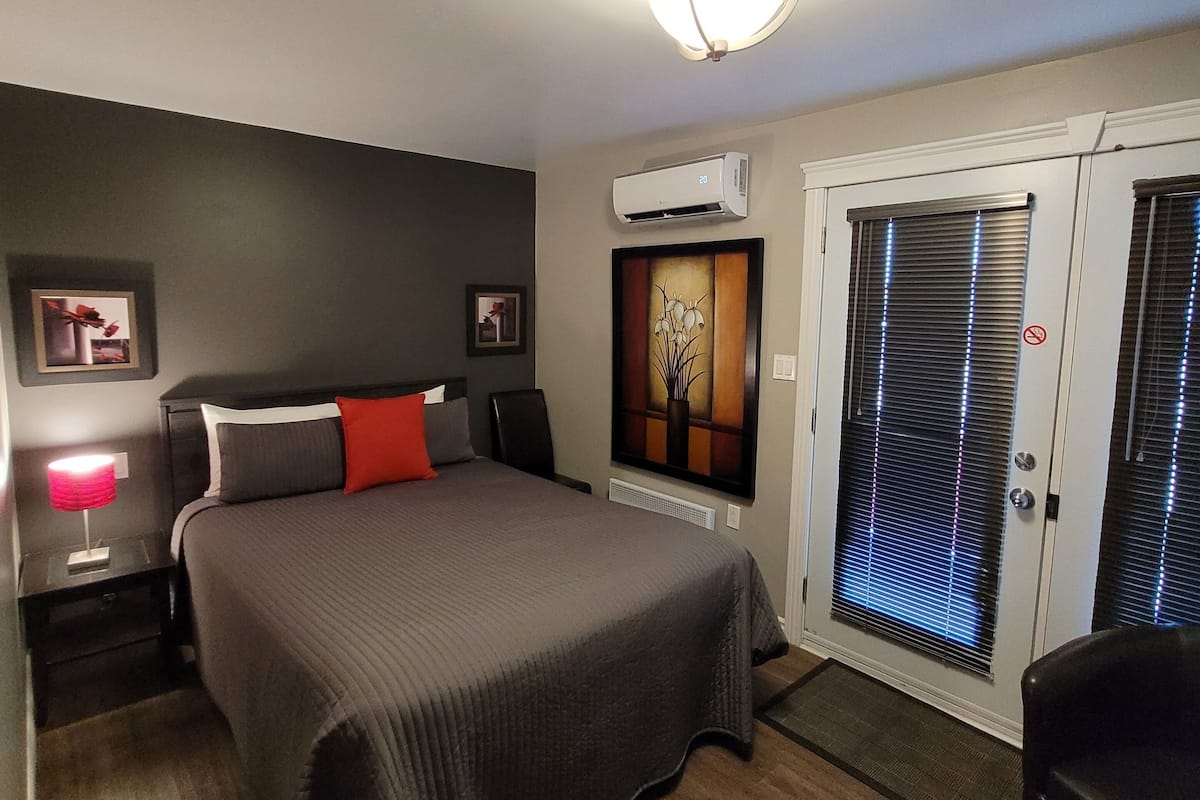 Una habitación de hotel con cama, mesita de noche con lámpara, unidad de aire acondicionado y puerta con persianas.