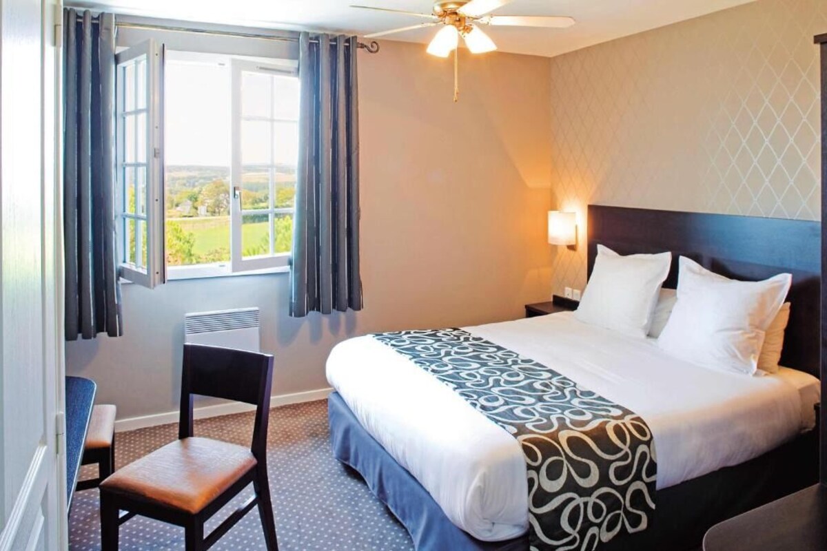 A hotel room with a bed, a chair, a window with curtains, a ceiling fan, and a wall-mounted lamp.