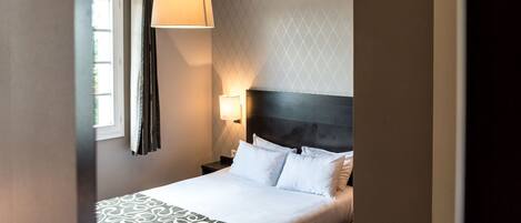 A hotel room with a bed, a nightstand, a wall-mounted lamp, and a window with curtains.