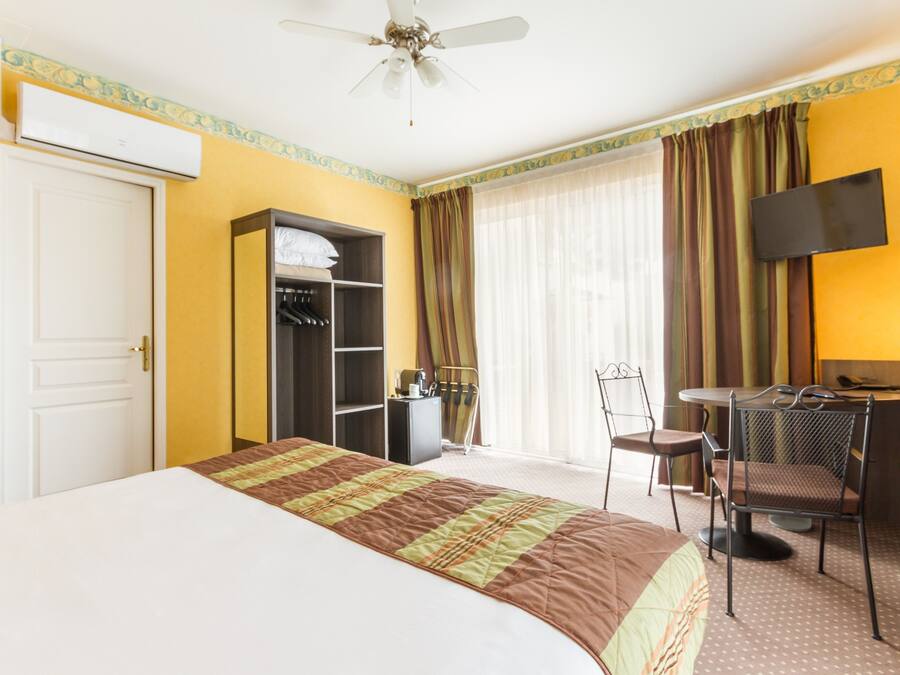 Habitació d'hotel amb un llit, un armari, una taula de nit amb cadira, una televisió i un ventilador de sostre.