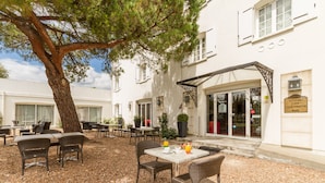 An outdoor patio with wicker furniture, a table with a drink, and a tree providing shade.