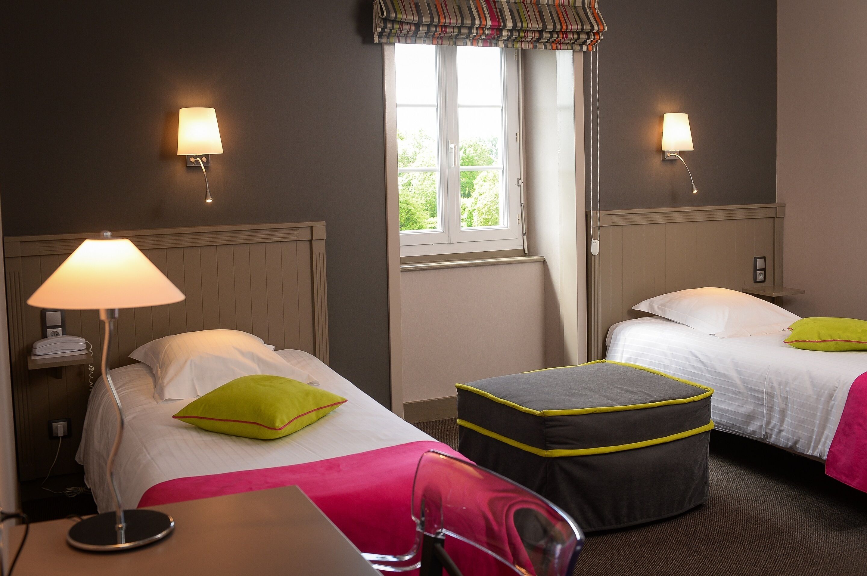 Een hotelkamer met twee bedden, elk voorzien van een kussen en een bedlampje. Er is een raam met gordijn, en een stoel is in de voorgrond zichtbaar.