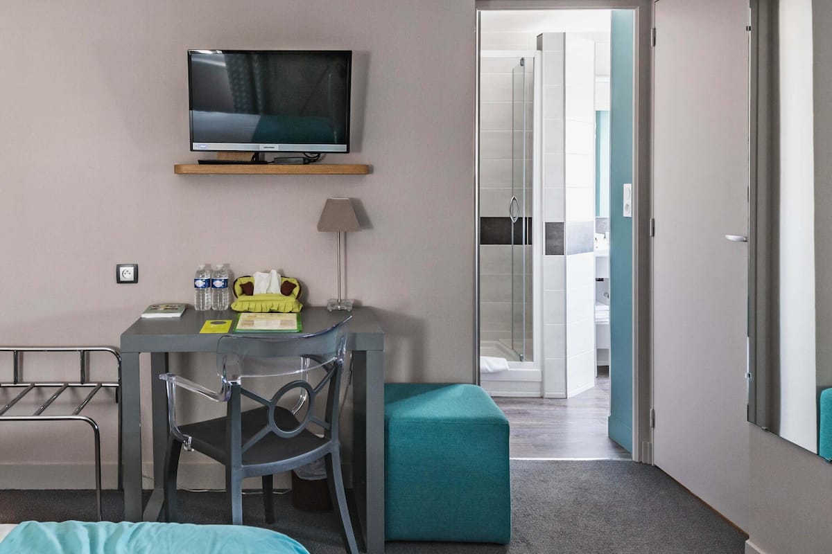 Hotellihuone, jossa on sänky, työpöytä tuolilla, televisio, kylpyhuone, joka näkyy avoimesta ovesta, ja pieni ikkuna.