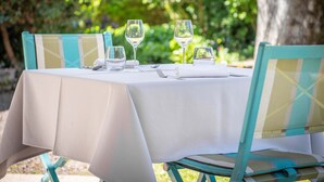 Mesa posta ao ar livre com toalha branca, duas cadeiras com almofadas xadrez e copos de vinho.