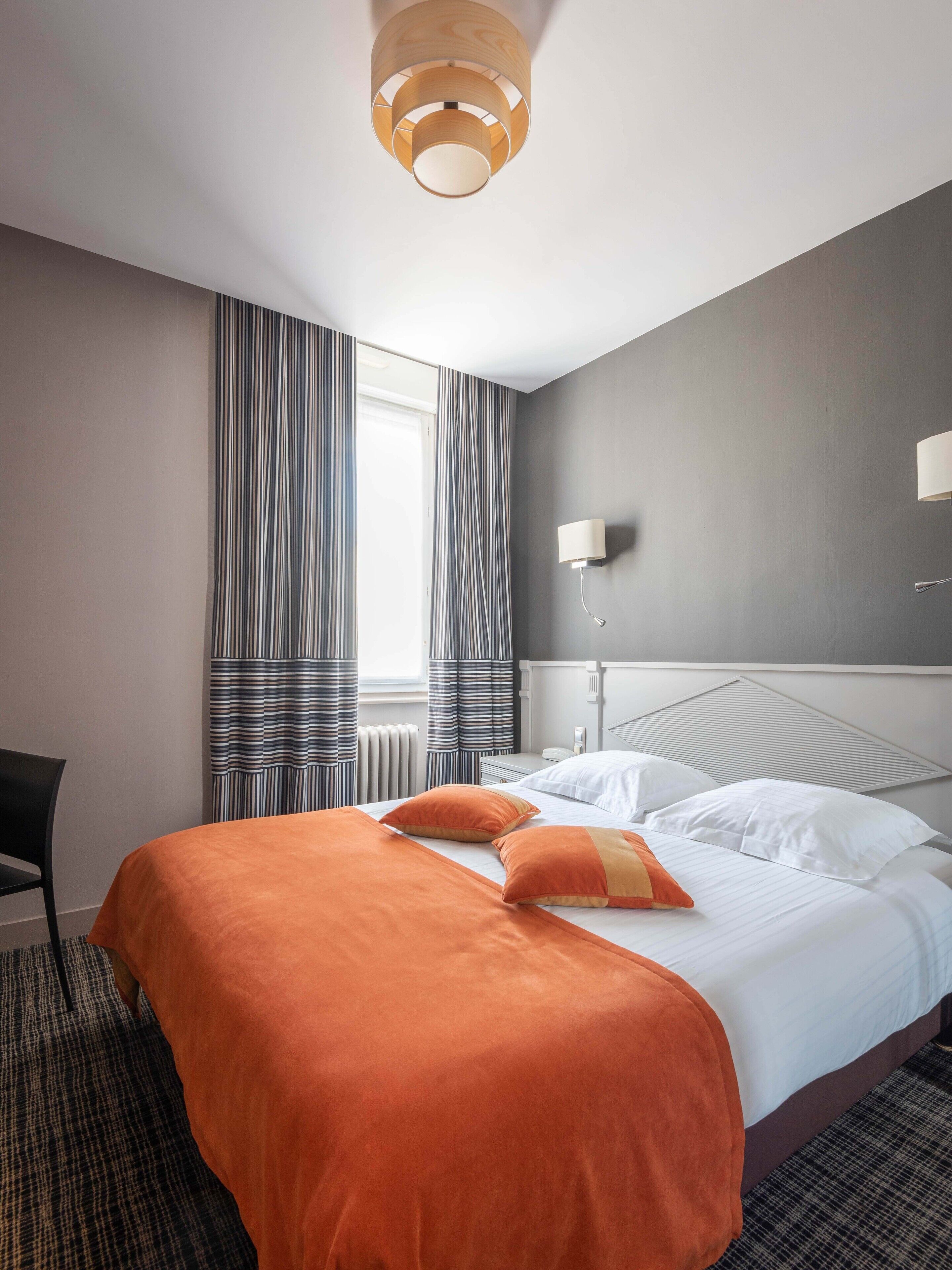 A hotel room with a large bed, striped curtains, a modern light fixture, and a wall-mounted lamp.