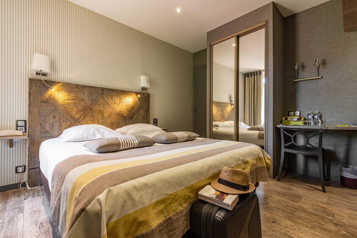 Hotellihuone, jossa on suuri sänky, yöpöytä, peili, työpöytä ja matkalaukku.