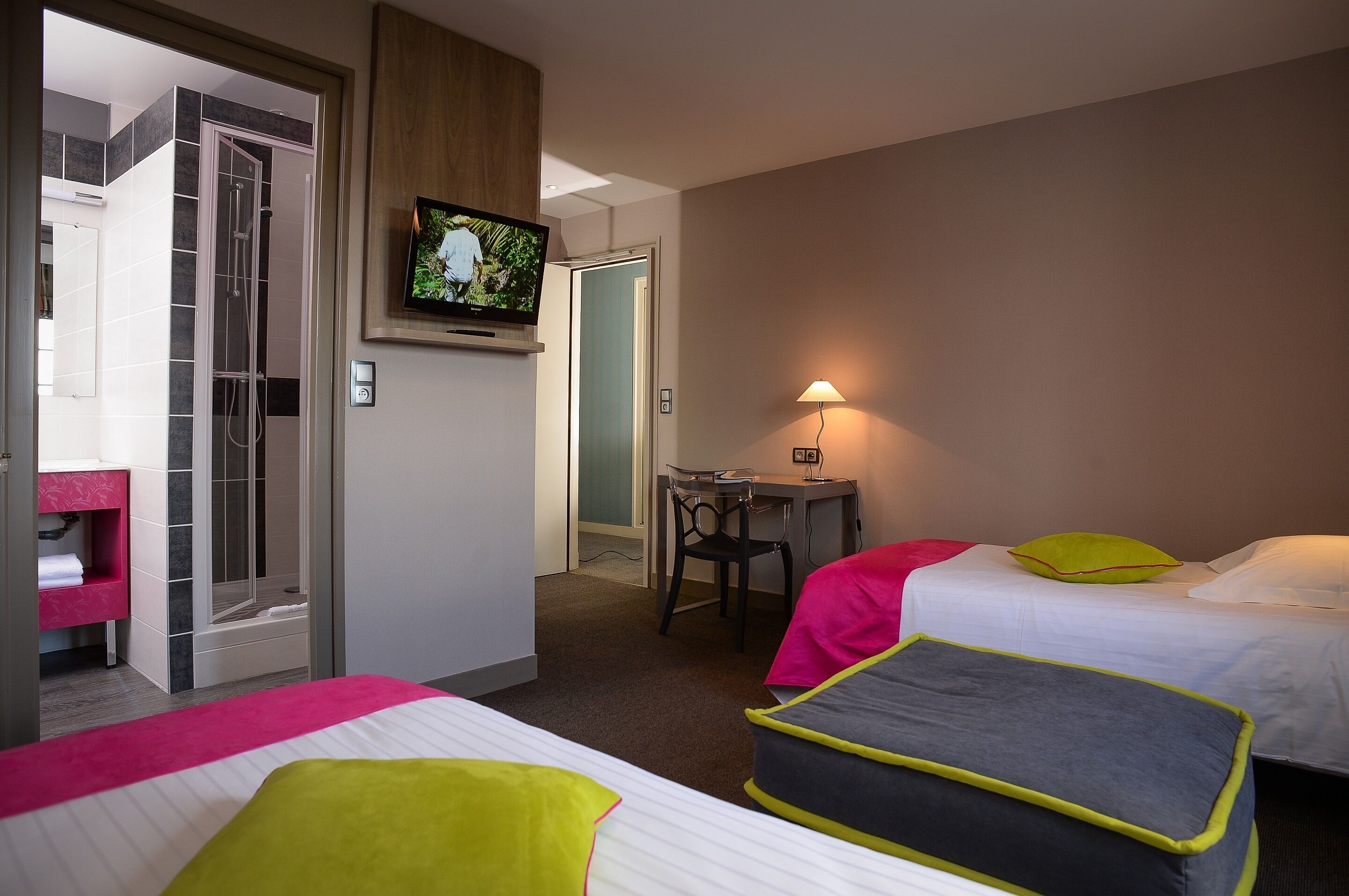 A hotel room with two beds, a desk, a TV, and a shower area.