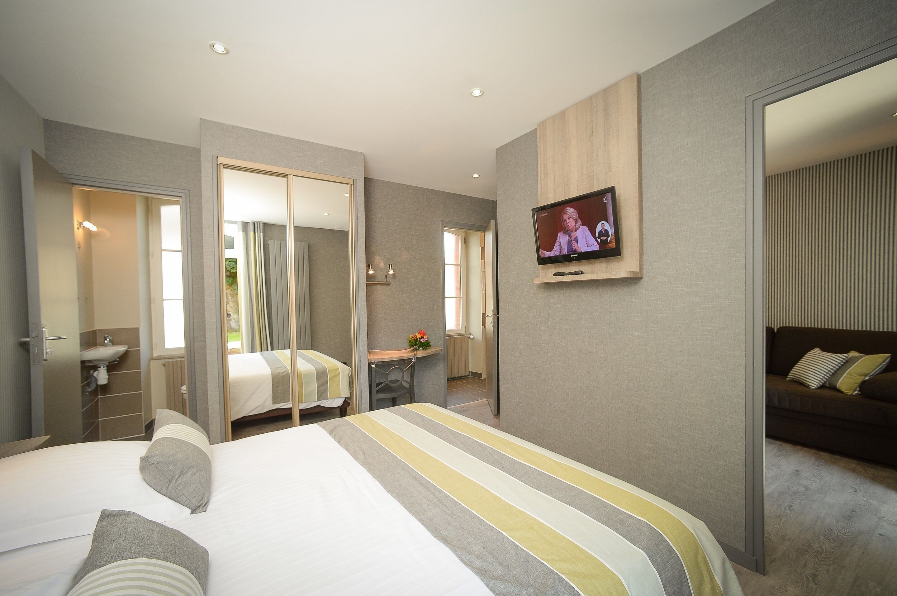 A modern hotel room with a large bed, a TV mounted on the wall, a mirror, and a small desk with a chair.