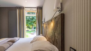 Quarto de hotel com uma cama grande, cabeceira de madeira, mesa de cabeceira e vista para um jardim através de uma porta de vidro.