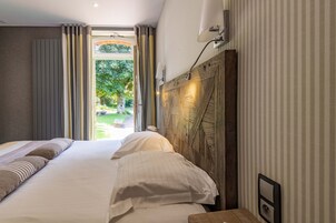 Habitación de hotel con una cama grande, cabecera de madera, mesita de noche y vistas a un jardín a través de una puerta de cristal.