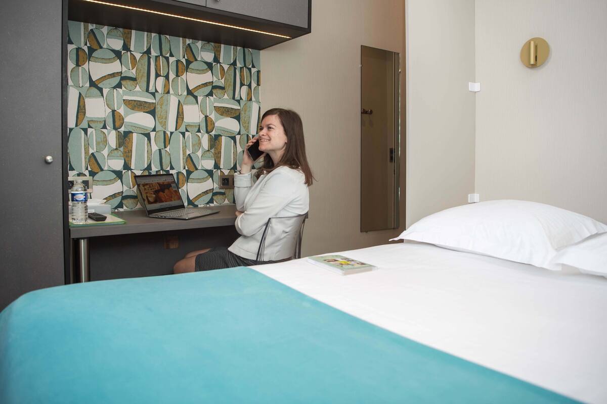 Hotellihuone, jossa on sänky, työpöytä kannettavalla tietokoneella, tuoli, geometrisesti kuvioitu seinä ja ovi.
