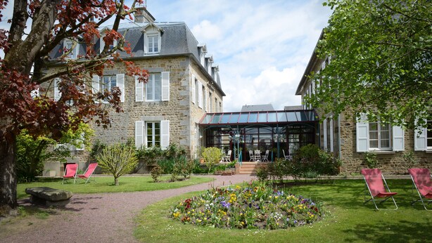 Un bâtiment en pierre doté d’une véranda en verre, un jardin fleuri et des fauteuils rouges.