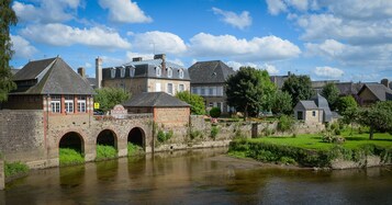 Un puente de piedra histórico sobre un río tranquilo, que conduce a una hilera de casas tradicionales europeas con ventanas arqueadas y una mezcla de piedra y ladrillo.