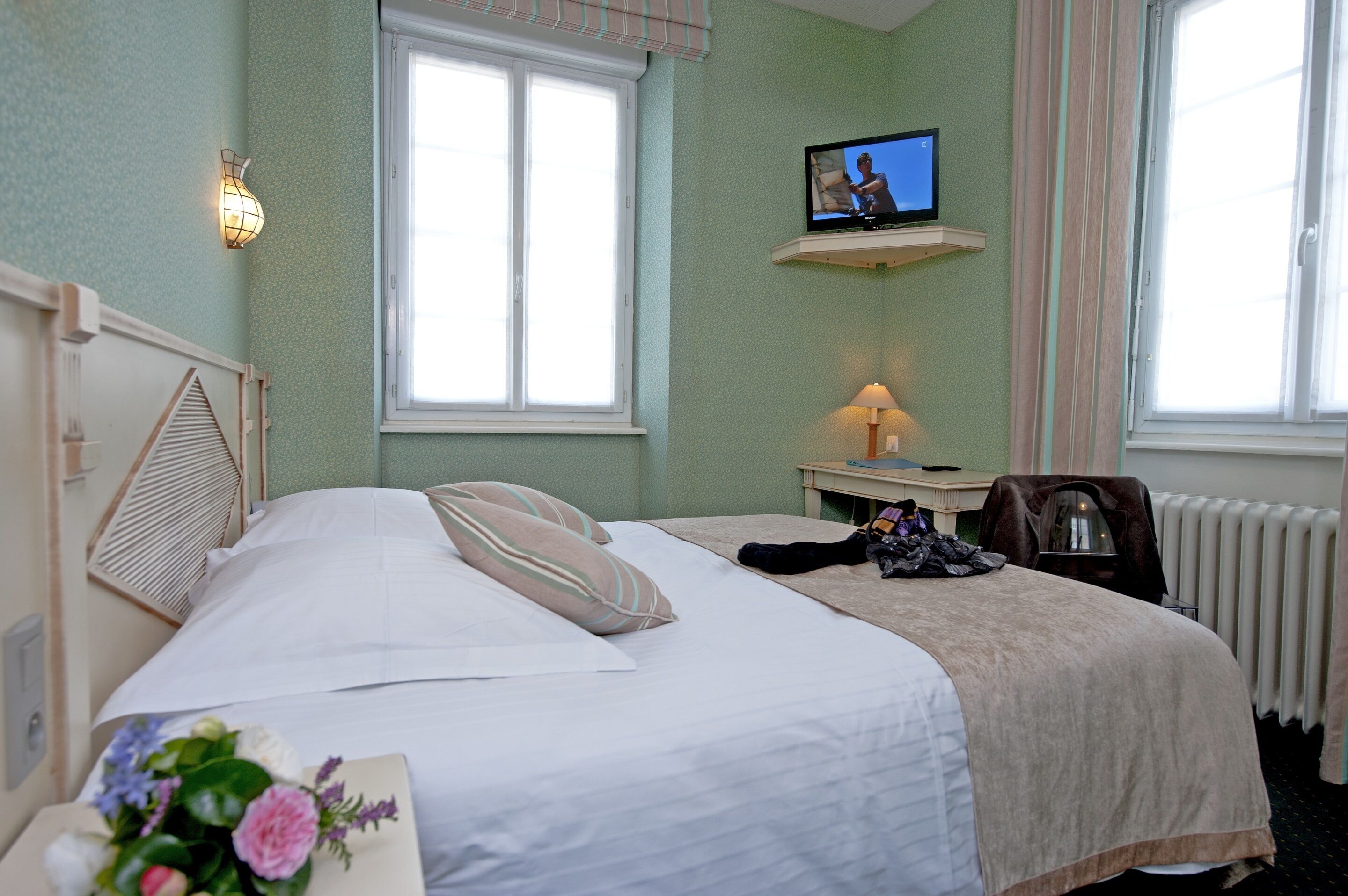 Une chambre d’hôtel avec un lit, une table de chevet avec une lampe, un téléviseur posé sur une étagère et une fenêtre avec des rideaux.