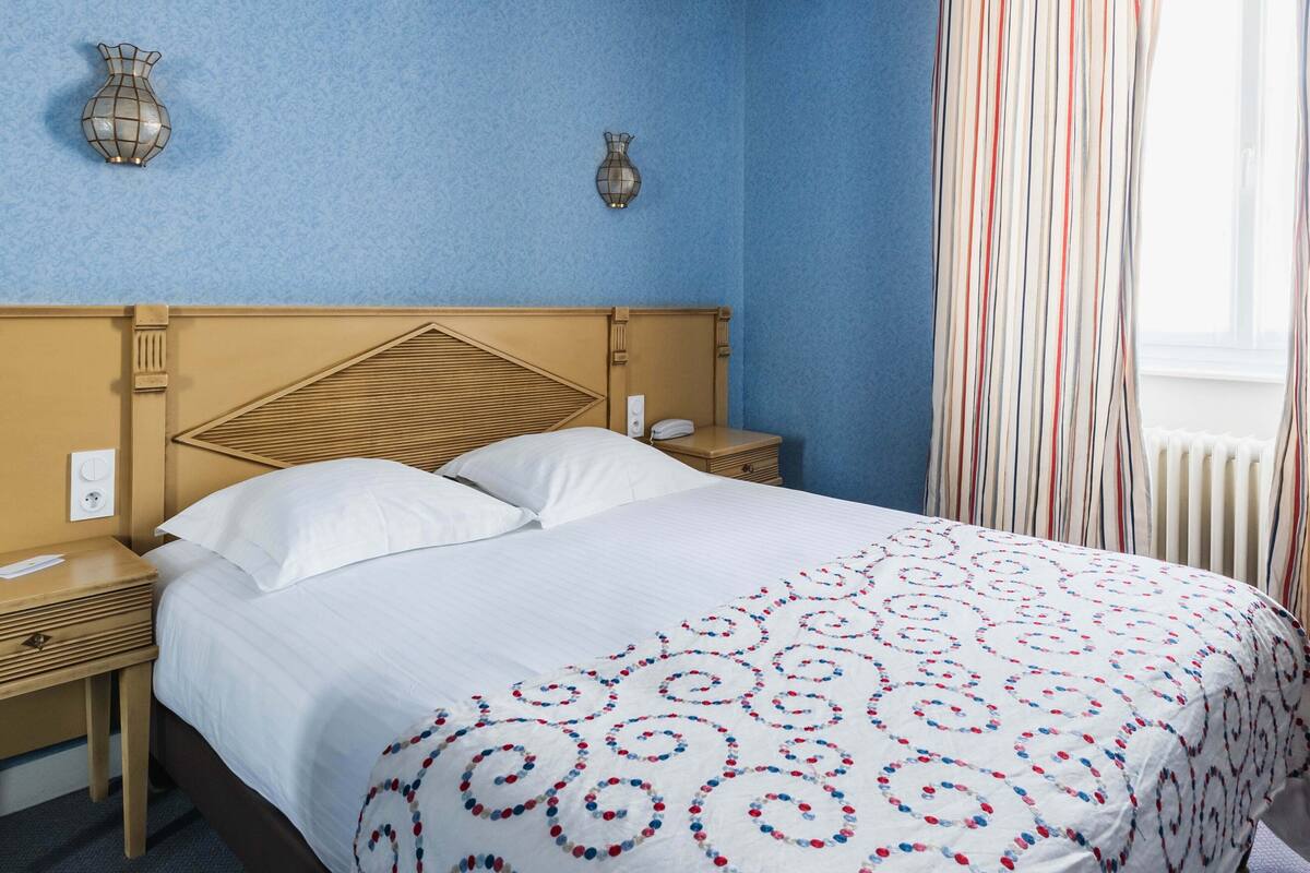 Hotellihuone, jossa on sänky, yöpöytä, seinään kiinnitetty lamppu ja ikkuna, jossa on verhot.
