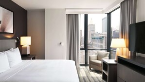 Habitación de hotel con una cama grande, una silla, un televisor y una ventana con vista a la ciudad.