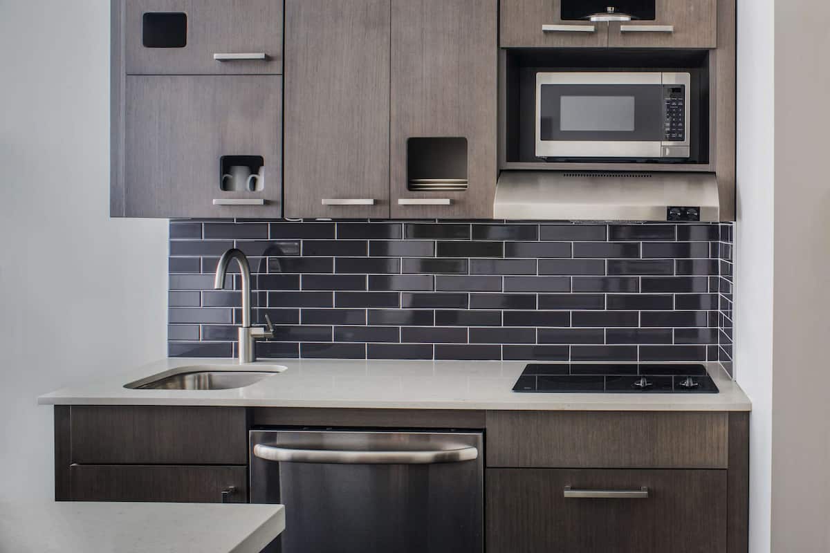 A modern kitchen with dark wood cabinets, a stainless steel dishwasher, a microwave, and a black tiled backsplash.