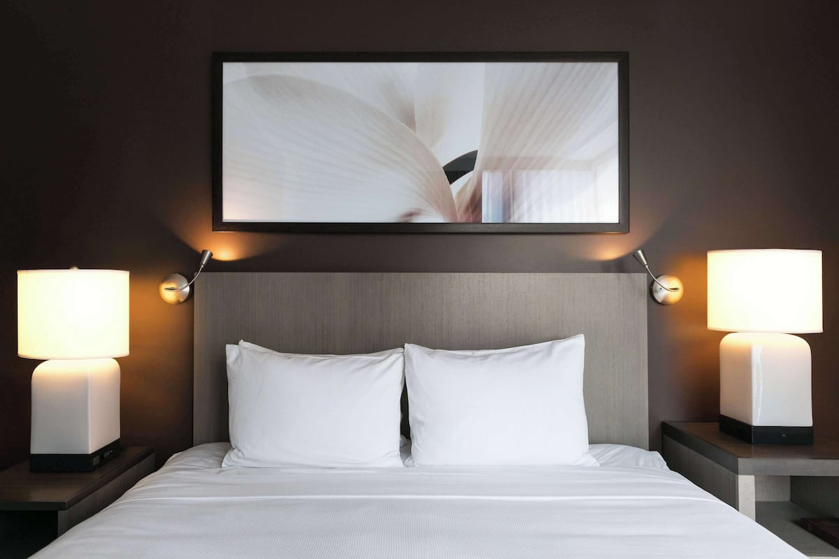 A hotel room with a neatly made bed, two bedside lamps, and a framed artwork above the bed.
