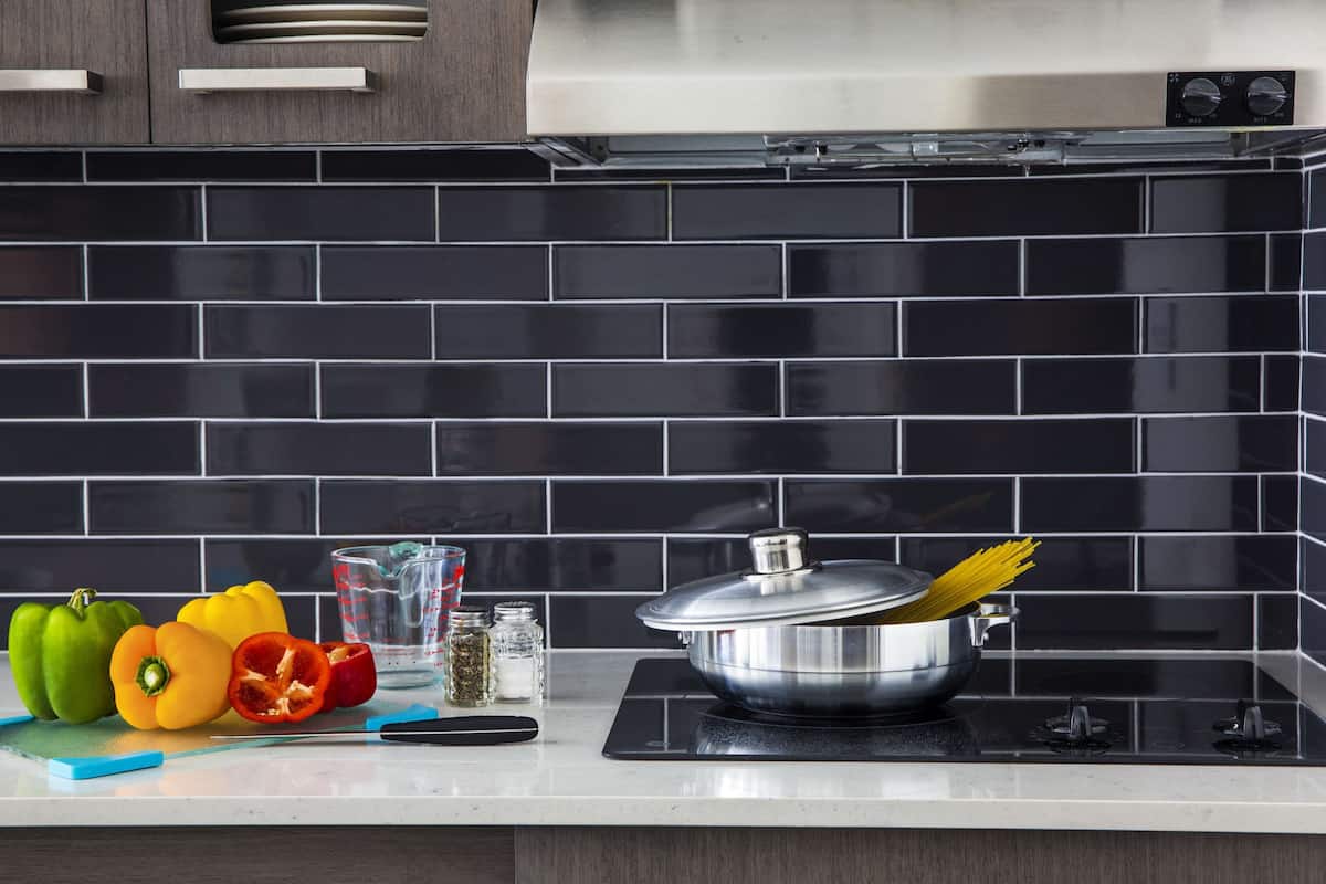 A kitchen with a black tiled backsplash, a stainless steel pot on an induction cooktop, and colorful bell peppers on the counter.