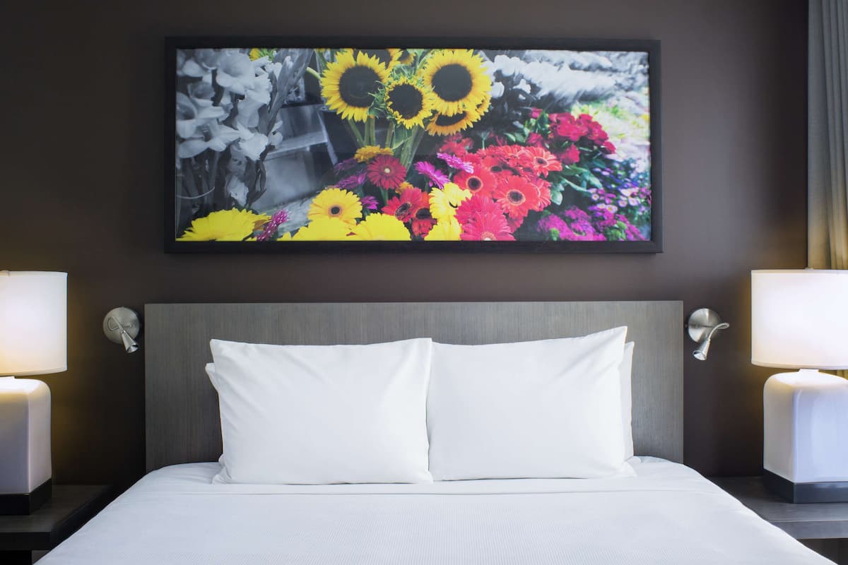A hotel room with a large bed, white bedding, two bedside lamps, and a framed floral artwork above the bed.