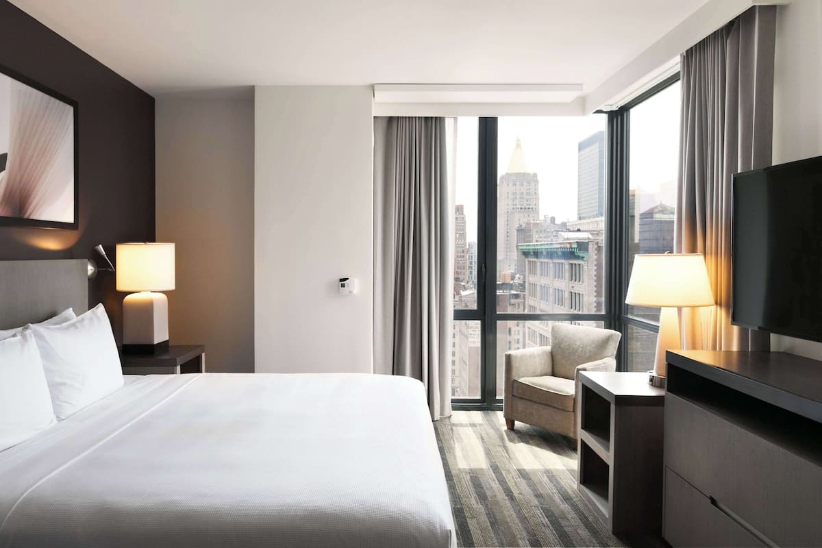 A hotel room with a large bed, two bedside tables with lamps, a TV, a chair, and a view of a cityscape through a large window.