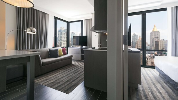Una habitación de hotel moderna con sofá, mesa de comedor, kitchenette y un amplio ventanal con vistas a la ciudad.