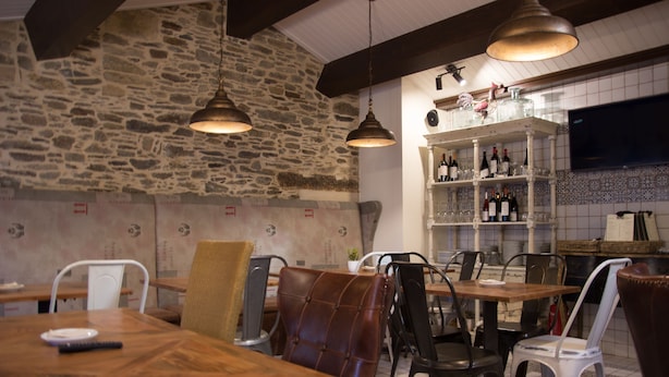 A restaurant interior with stone walls, hanging pendant lights, a bar area with shelves of bottles, and a television mounted on the wall. - La Casona de Nené (Arzua)