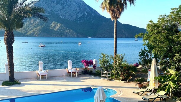 Zona de piscina con sillas de descanso y sombrillas, con vistas a una masa de agua con barcos y montañas al fondo.