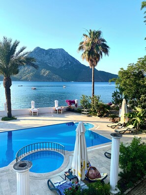 A pool area with lounge chairs and umbrellas, overlooking a body of water with boats and mountains in the background. - Ford Hotel - Special Class (Kumluca)