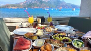 Una mesa de desayuno con varios platos, vista al mar con barcos y una montaña al fondo.