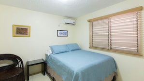 Une chambre avec un lit, une petite table, une chaise, une fenêtre avec des stores et un climatiseur fixé au mur.