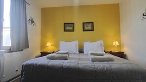 Una cama bien hecha con dos almohadas y dos toallas dobladas, una mesita de noche con una lámpara, y dos cuadros en la pared.