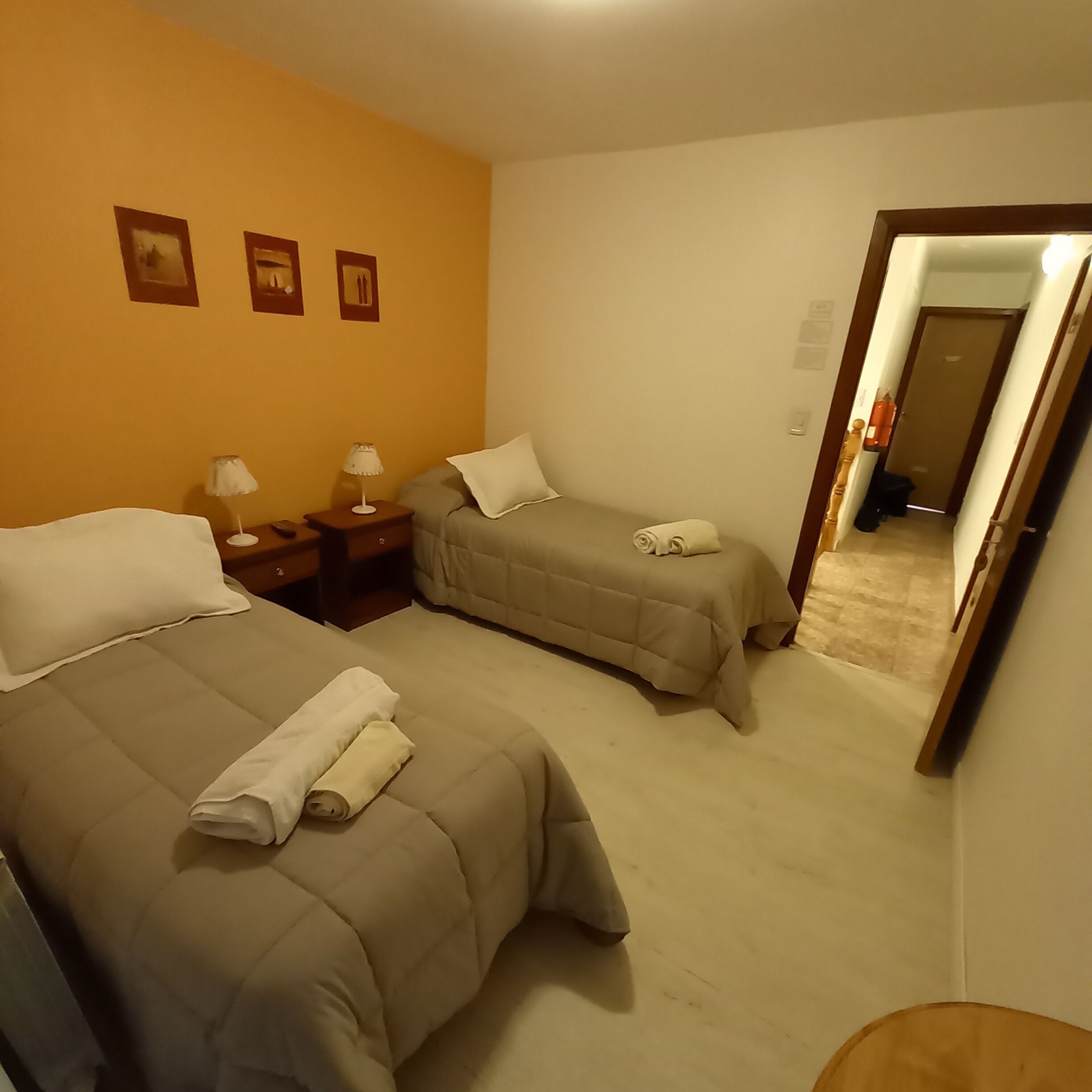A hotel room with two beds, each with a pillow and a rolled towel. There are two bedside tables with lamps. The walls are painted yellow, and there are three framed pictures on the wall.