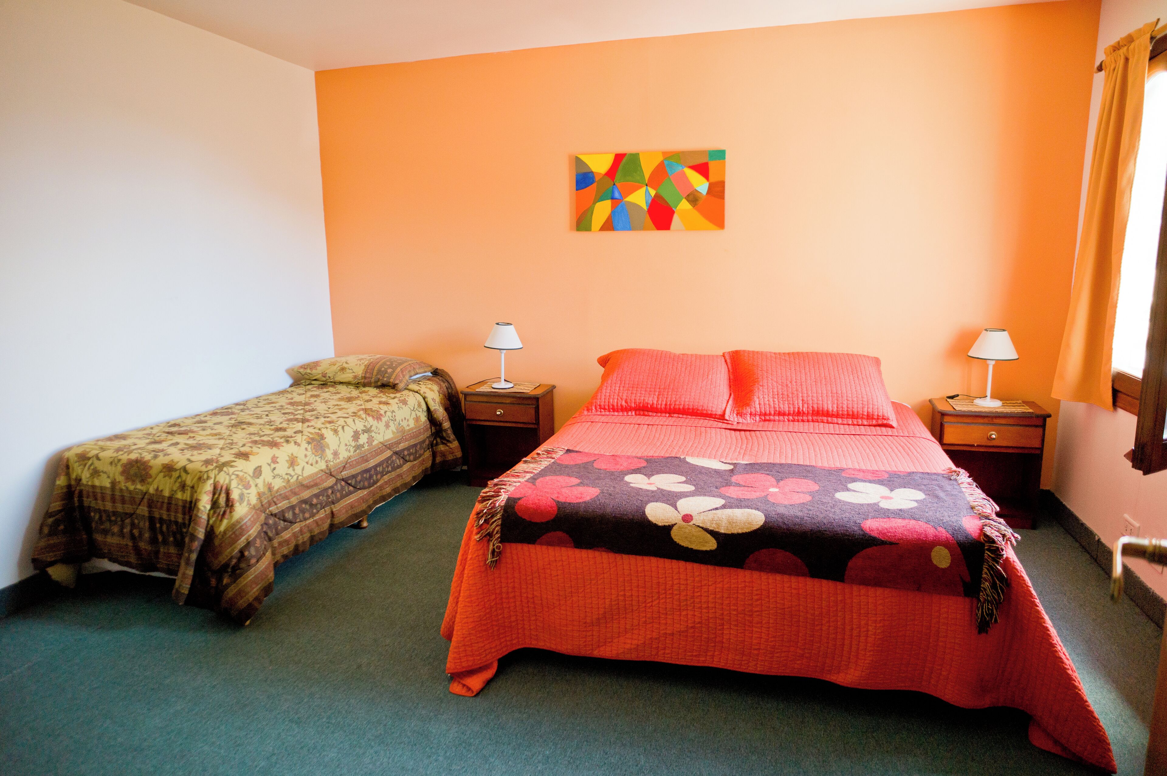 A room with two beds, each with a different style of bedding. There are two bedside tables with lamps, a colorful abstract painting on the wall, and a window with curtains.