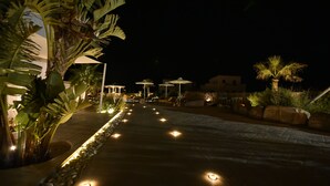 Un sendero nocturno con luces iluminadas, palmeras y sombrillas de exterior.