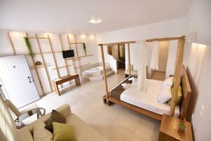 Quarto moderno com cama de dossel, sofá, televisão e banheiro ao fundo.