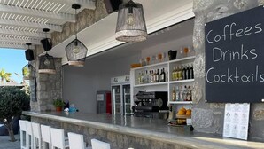 Zona de bar al aire libre con taburetes de mimbre blanco, pared de piedra y un menú en pizarra que ofrece cafés, bebidas y cócteles.