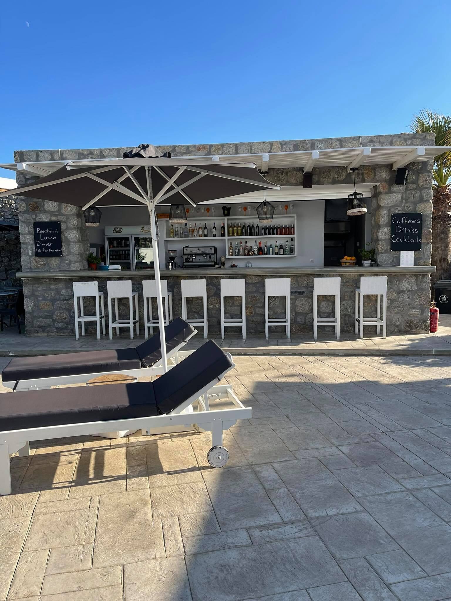 Un edificio de piedra con una zona de bar, mesas al aire libre con tumbonas y un cartel que anuncia desayuno, comida, cena y cócteles.