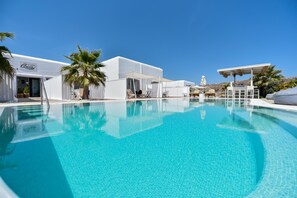 A swimming pool with clear blue water, surrounded by white buildings and palm trees. - Queen Suites (Mykonos)