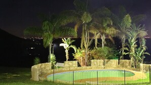 A nighttime view of a landscaped area with a swimming pool, palm trees, and a stone wall. - The Cradle Rest (Muldersdrift)