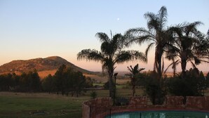 A swimming pool surrounded by a stone wall, with palm trees and a hilly landscape in the background. - The Cradle Rest (Muldersdrift)