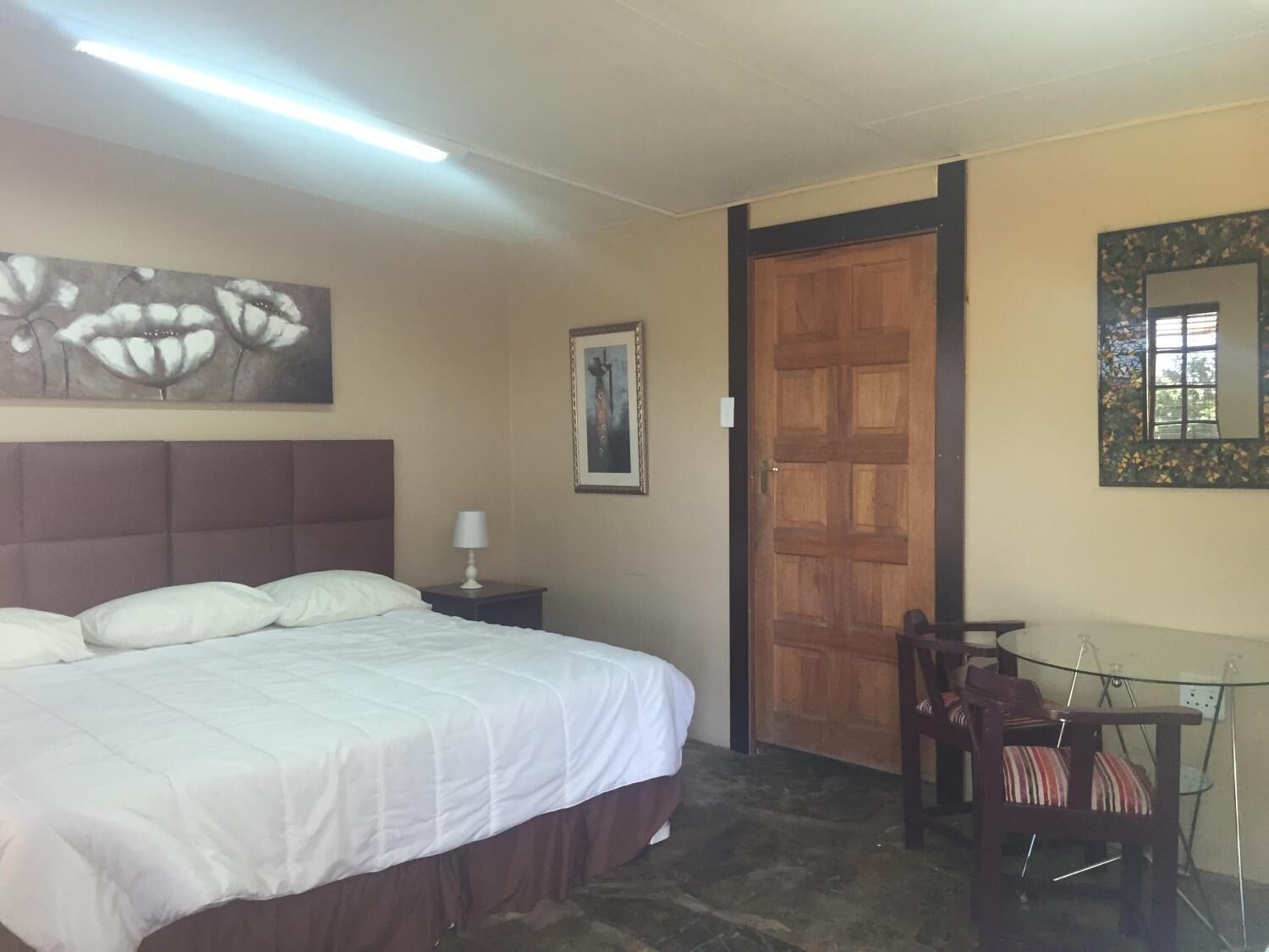 A hotel room with a bed, a nightstand, a chair, a table, a door, and a wall with artwork.