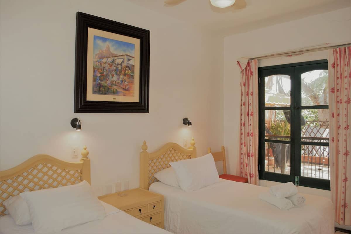 A hotel room with two beds, a framed painting, bedside lamps, a window with curtains, and a view of the outside.