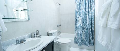 A modern bathroom with a white sink, toilet, and bathtub. There are glass shelves above the sink and a patterned shower curtain.