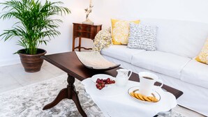 Una sala de estar con un sofá blanco, una mesa de centro con una bandeja de aperitivos, una planta en maceta y una lámpara.