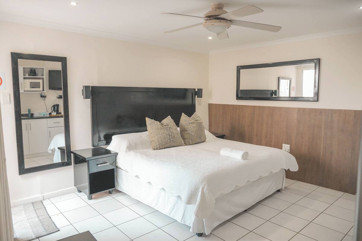 Une chambre avec un grand lit, une table de chevet, un ventilateur de plafond, un miroir et une vue sur un coin cuisine.