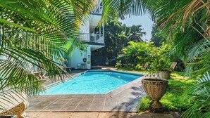 Una piscina rodeada de abundante vegetación y un edificio de dos pisos con balcones.
