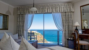 A hotel room with a large window offering a view of the ocean. There is a bed with pillows, a desk with a lamp, and a chair. - Headlands House Guest Lodge (Knysna)