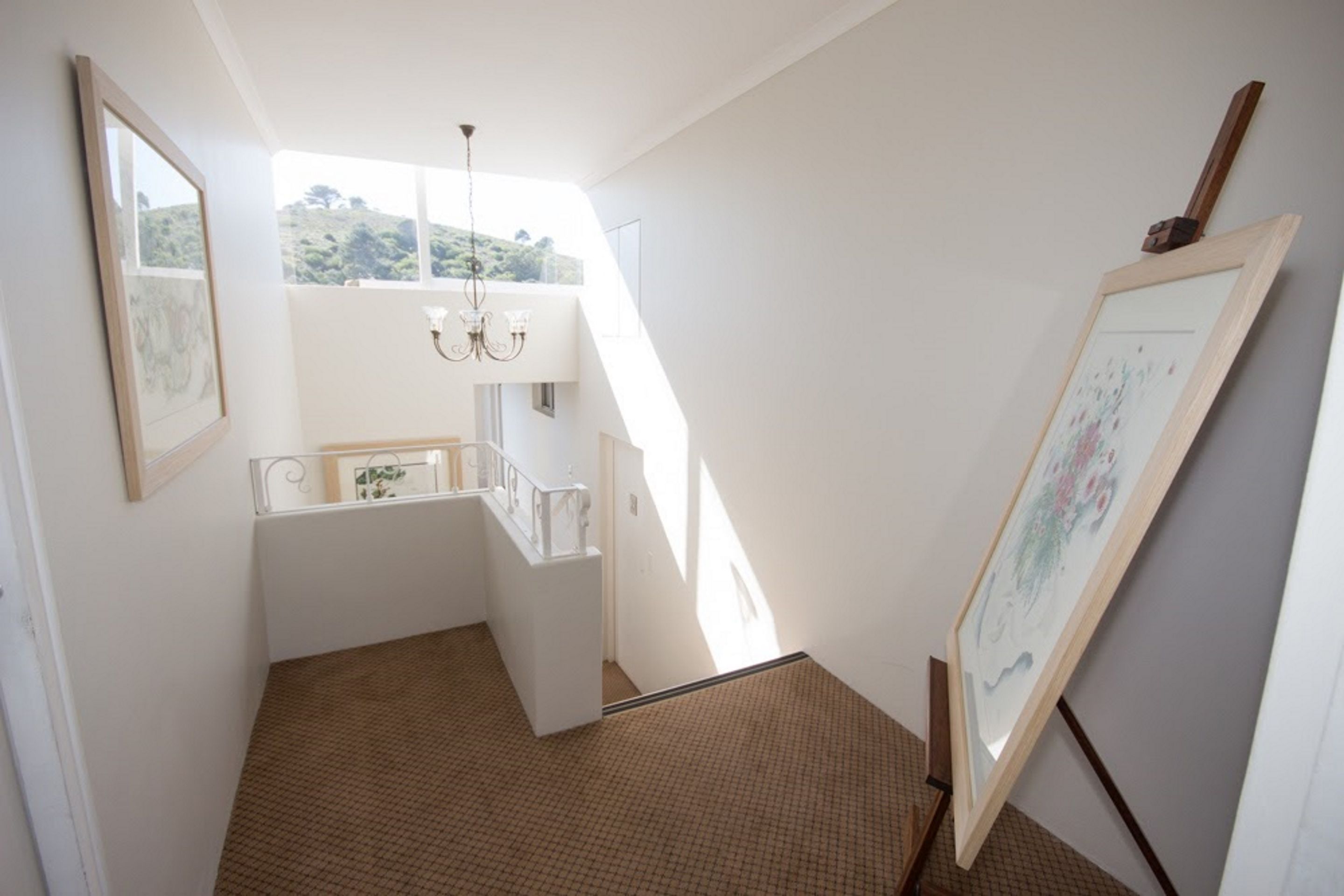 A narrow stairway with a glass railing, a chandelier, and framed artwork on the walls.