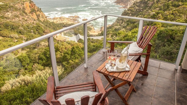 A balcony with a wooden table set for two, overlooking a coastal landscape with cliffs and the sea. - Headlands House Guest Lodge (Knysna)